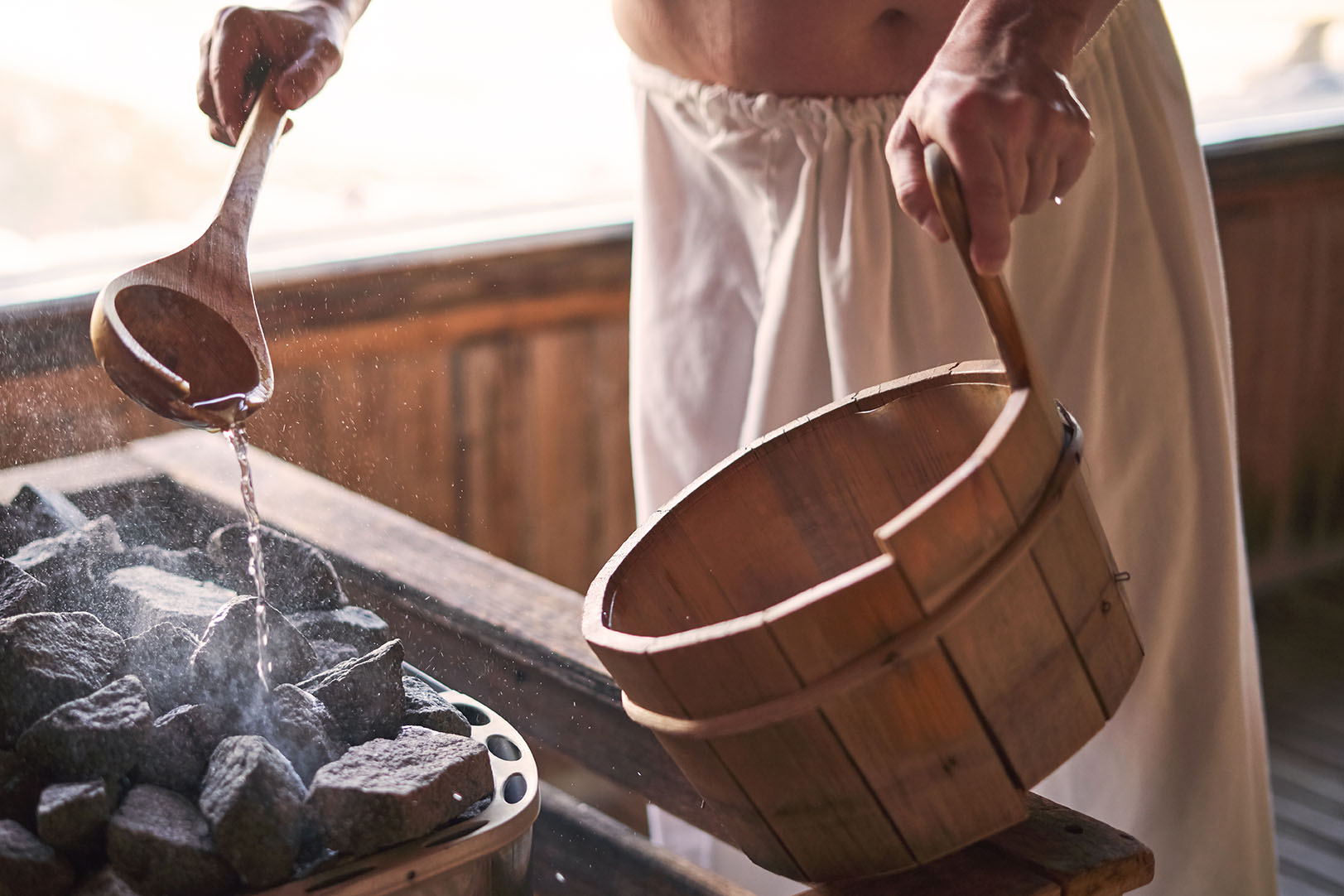 Man pouring water into hot stone in sauna room. Steam on the stones, spa and wellness concept, relax in hot finnish sauna.